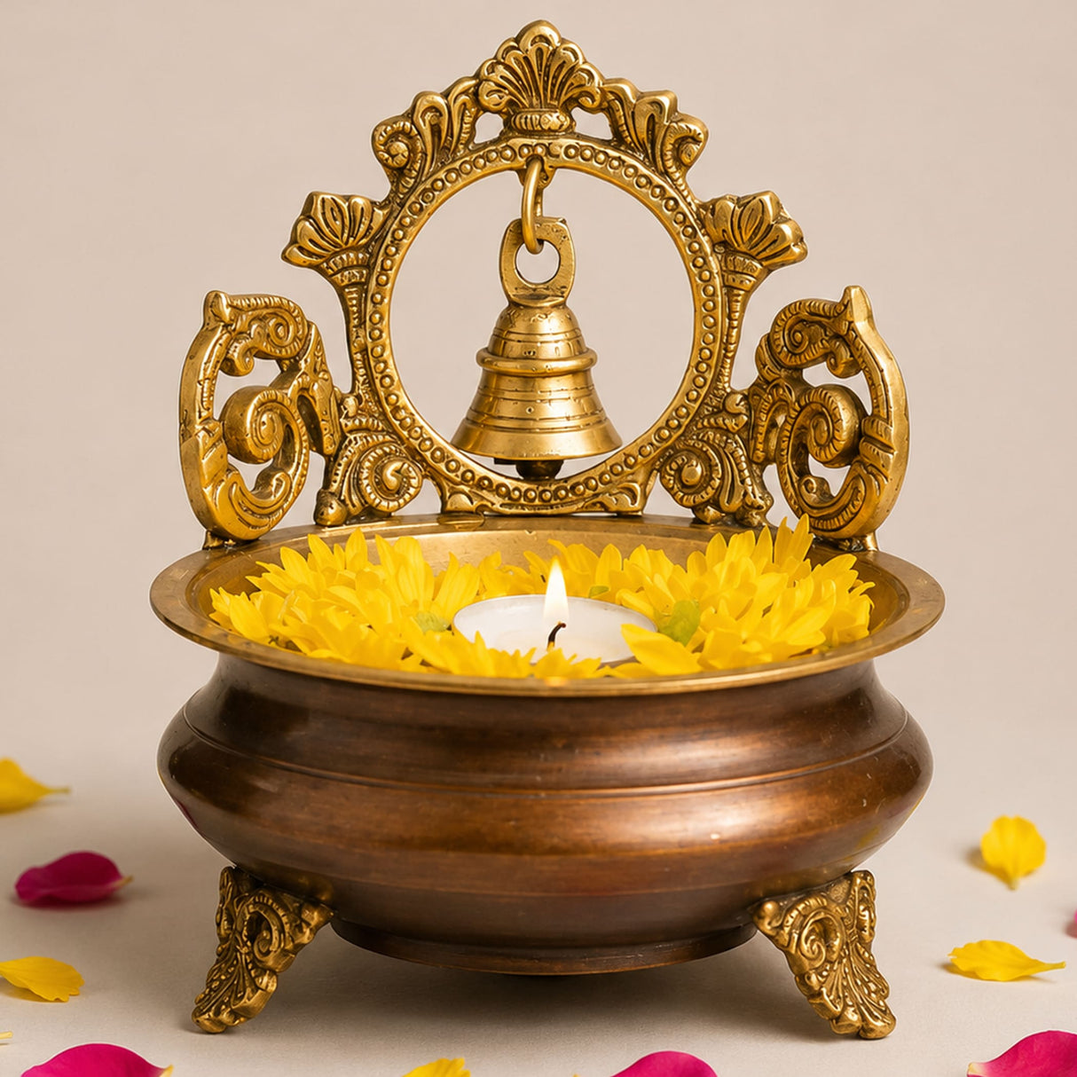 Golden ornate brass urli bowl with hanging bell and decorative handles featuring yellow marigold petals around lit candle