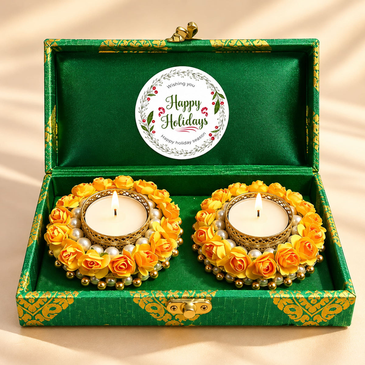 Happy holidays gift hamper with two ornate golden tealight candle holders adorned with yellow paper roses and white pearls inside a green satin-lined premium brocade box