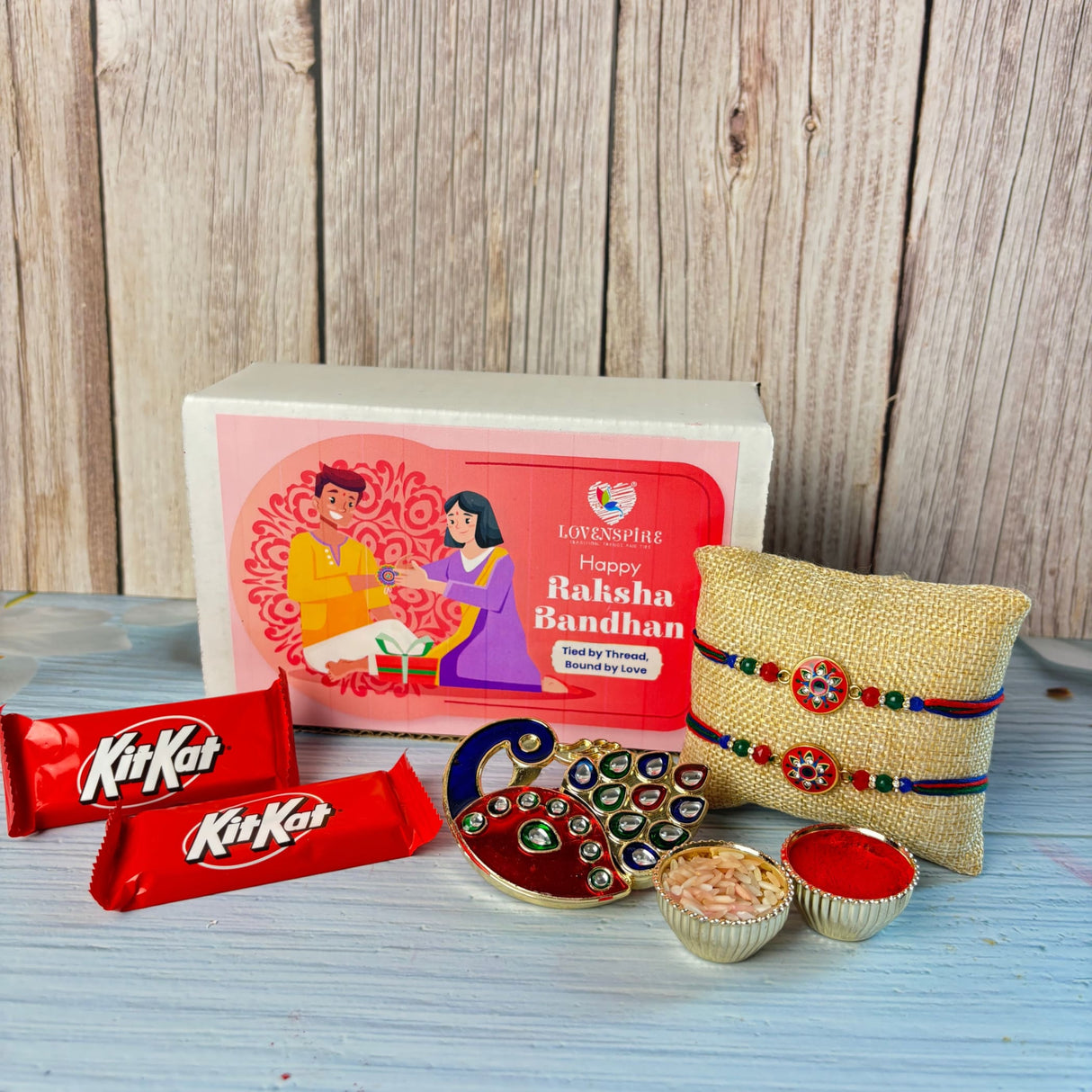 Gold-colored peacock-shaped designer rakhi with intricate red and green gemstones in gift hamper set for brother and bhabhi