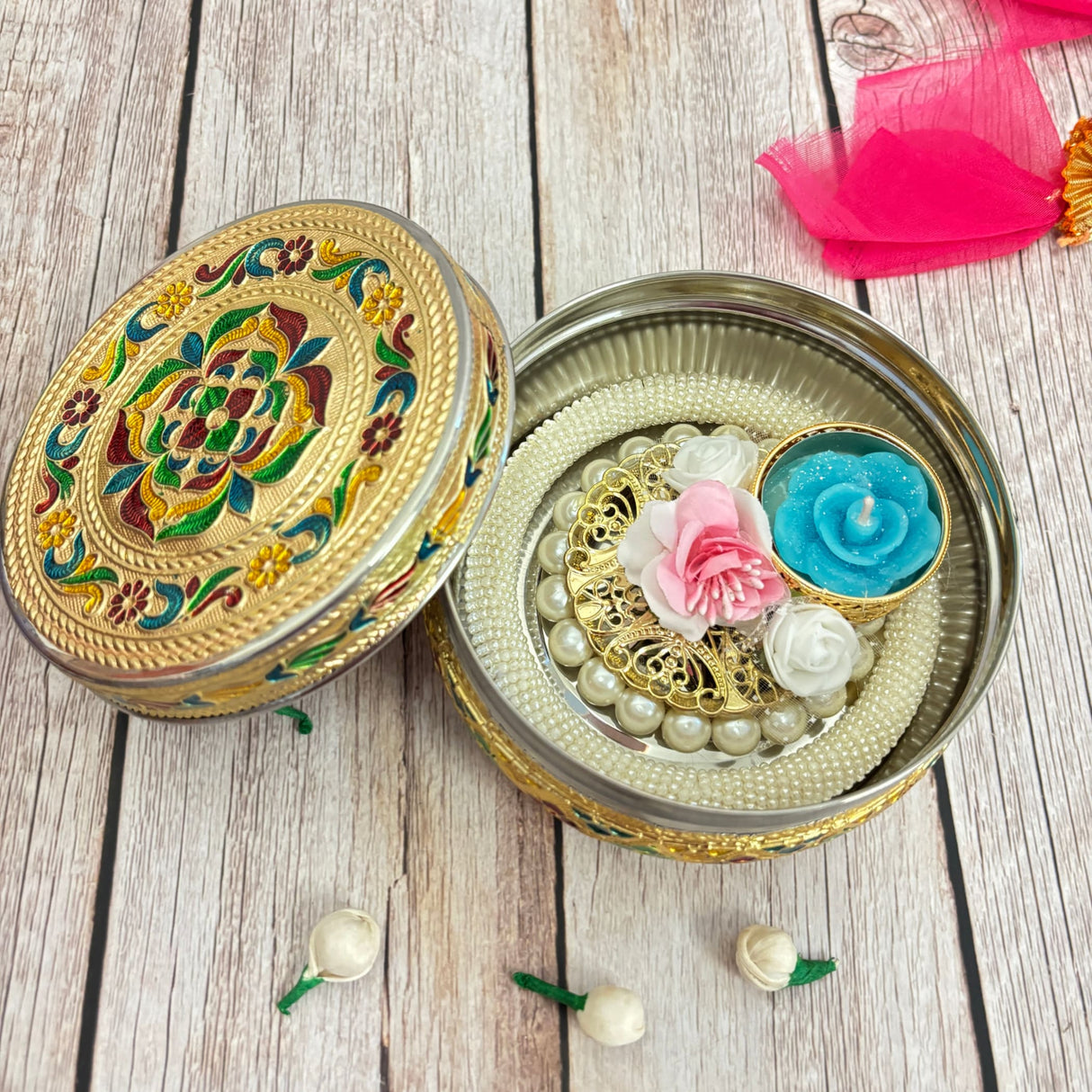 Open gold-colored metal box with enamel lid containing pearl necklace, decorative disk, flowers, and candle for Diwali gift hamper