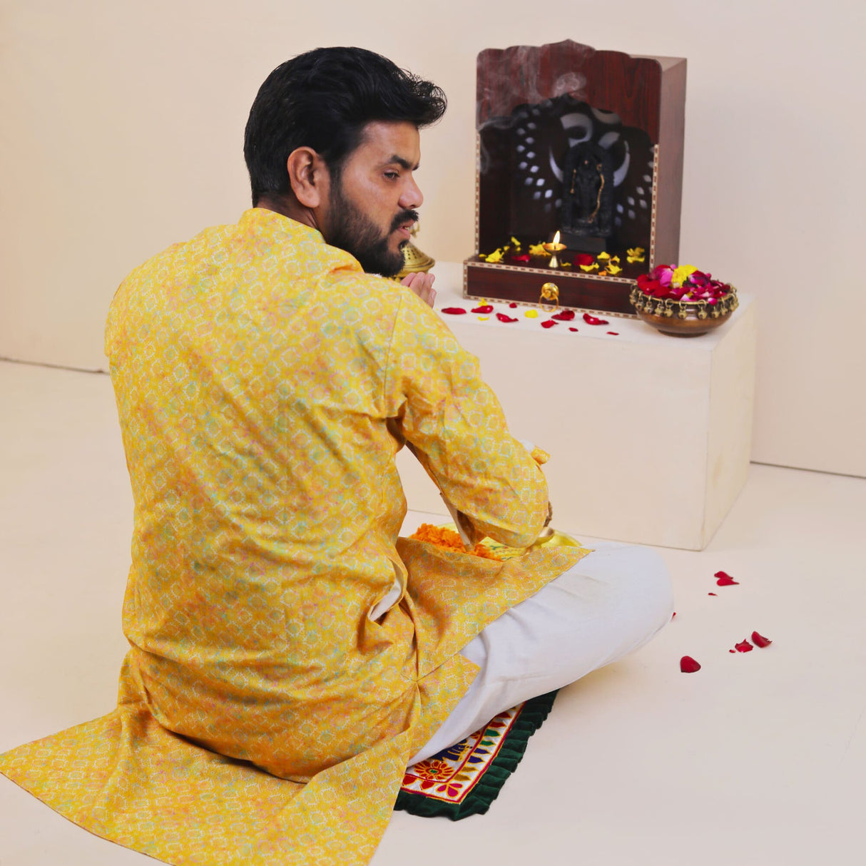 Man in yellow kurta sits on Elephant Pooja Aasan rug before ornate Ganesh shrine