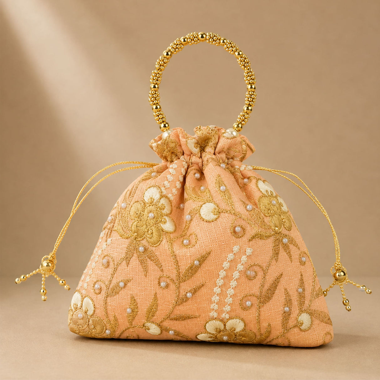 A peach-colored designer potli bag with embroidered gold floral details pearl embellishments and a beaded golden handle