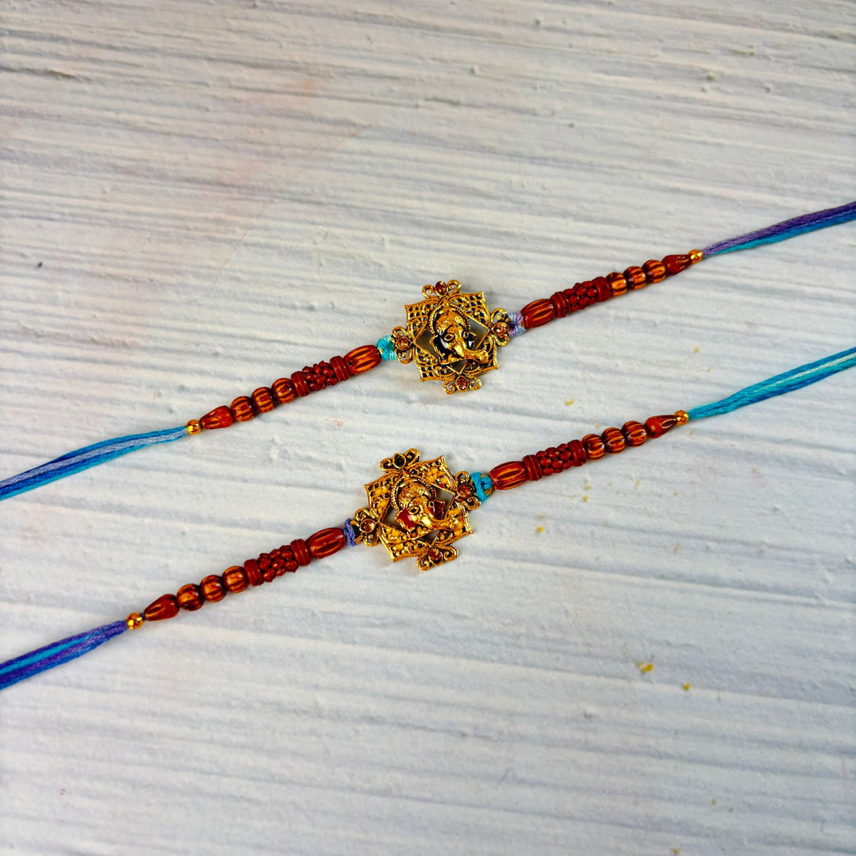Two designer ganesha rakhi bracelets with golden deity pendants on red and blue threads for raksha bandhan gift