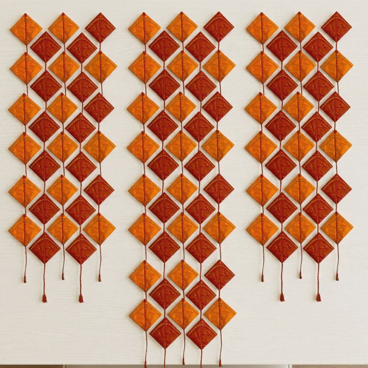 Orange and rust-colored diamond-shaped quilted wall hanging with stitched details and tassels for hindu wedding decor