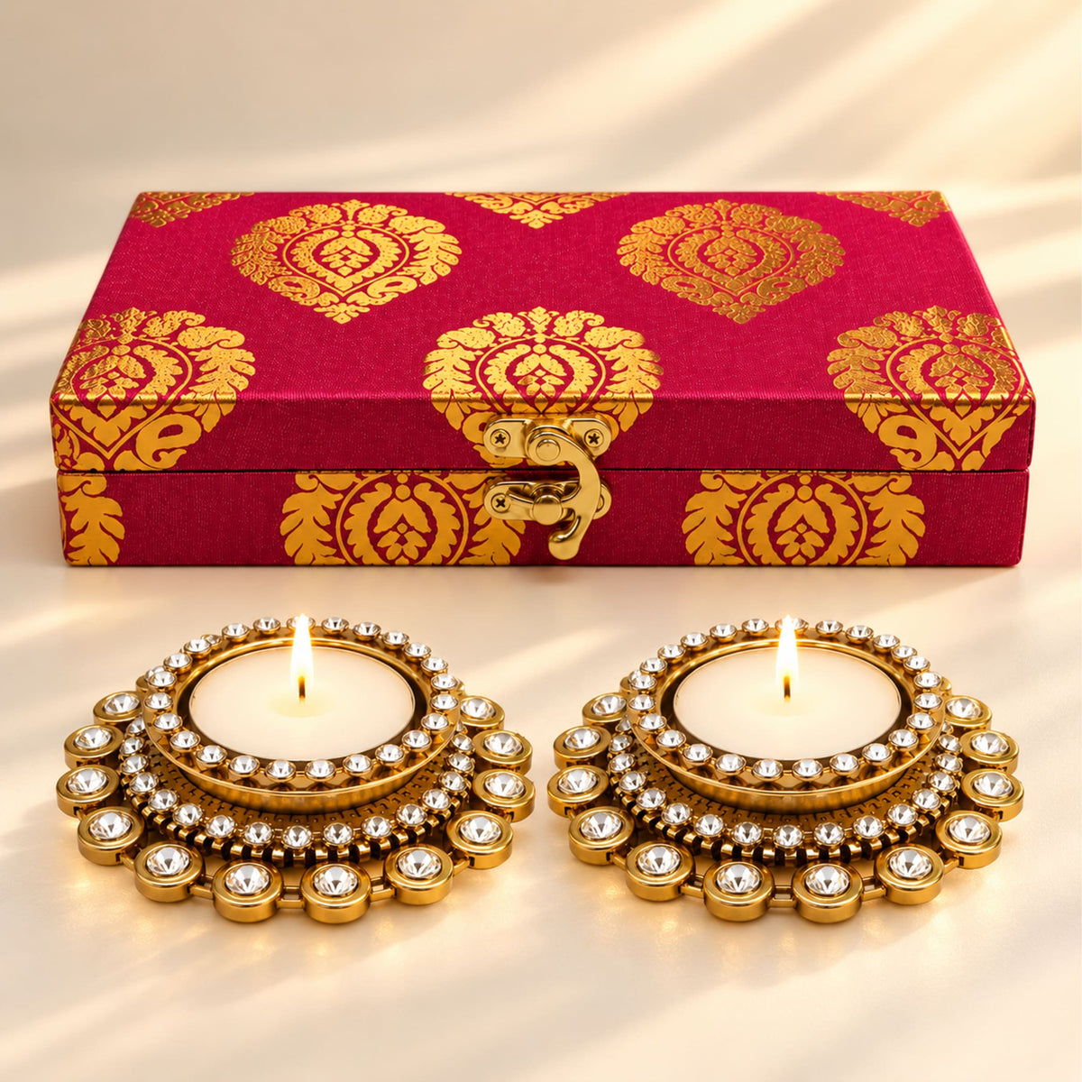 Gold-plated diya holders with sparkling crystals next to a red and gold patterned box for Diwali festival gifts