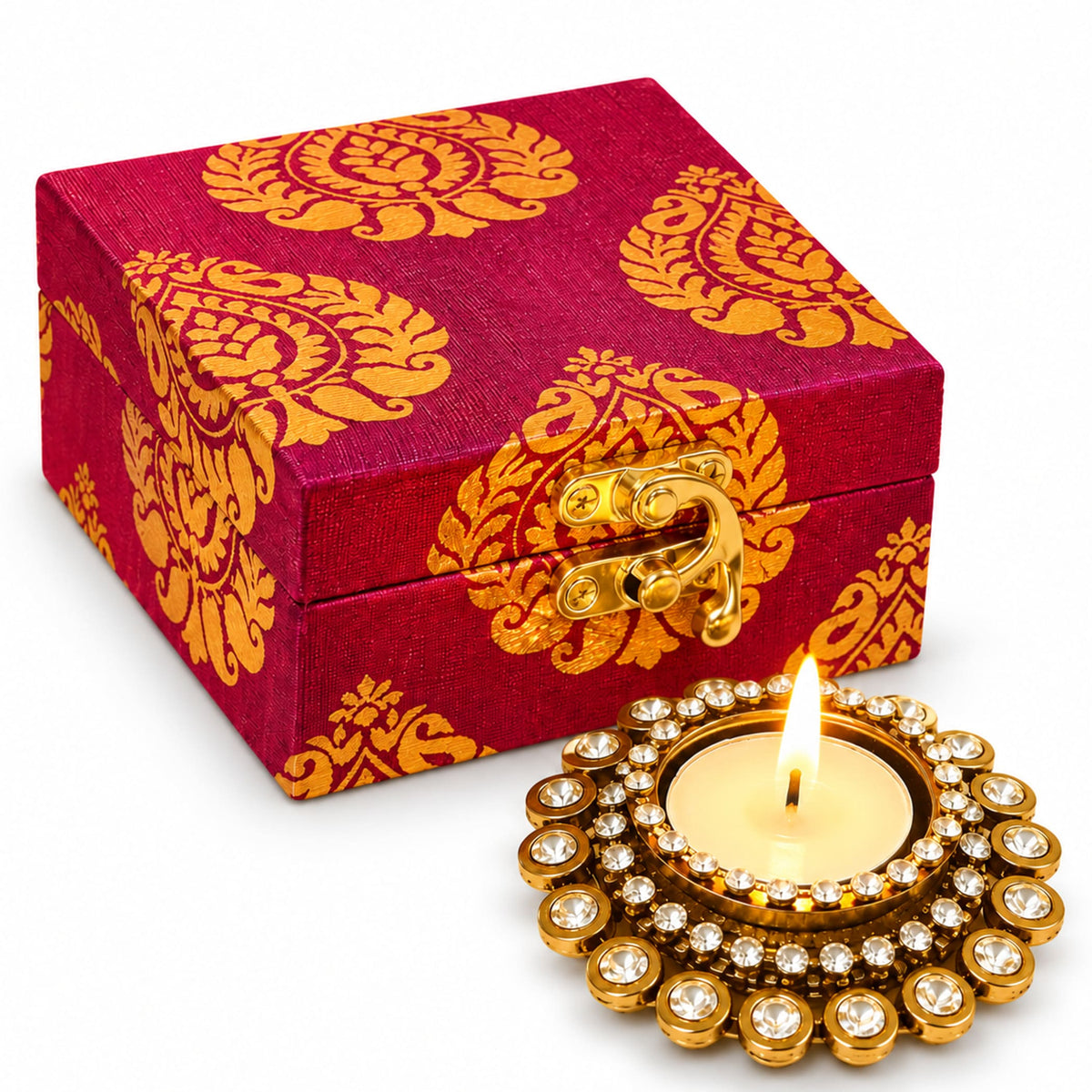 A maroon and gold patterned fabric box with golden clasp beside a diamond-encrusted brass diya candle holder for Diwali gifts