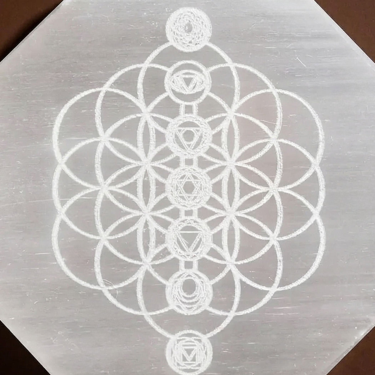 A translucent hexagonal selenite charging plate with white etched flower of life and seven chakra symbols for crystals