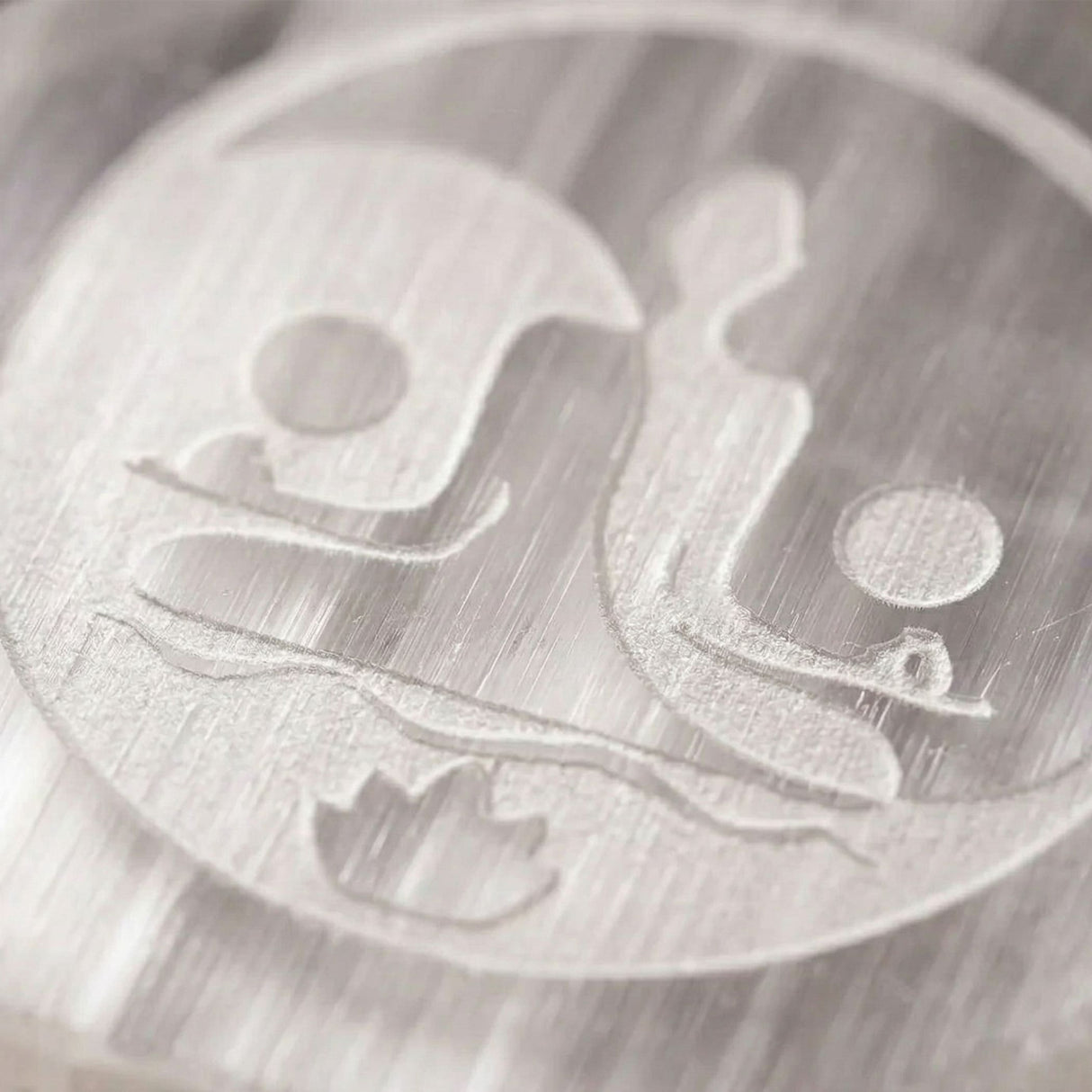 A circular brushed silver metal emblem with two stylized figures on a selenite charging plate for crystals