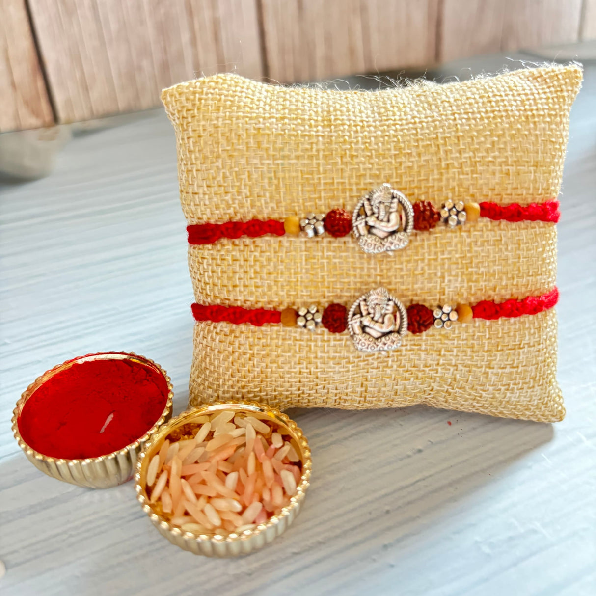 Designer silver tone ganesha rakhi set featuring two red and silver bracelets displayed on a beige jute pillow
