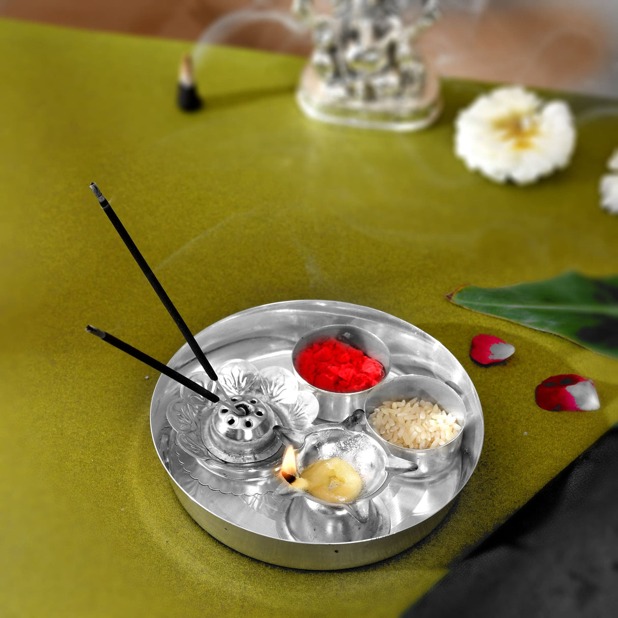Silver plated metallic pooja thali tray with three compartments holding red powder rice grains and a lit decorative diya
