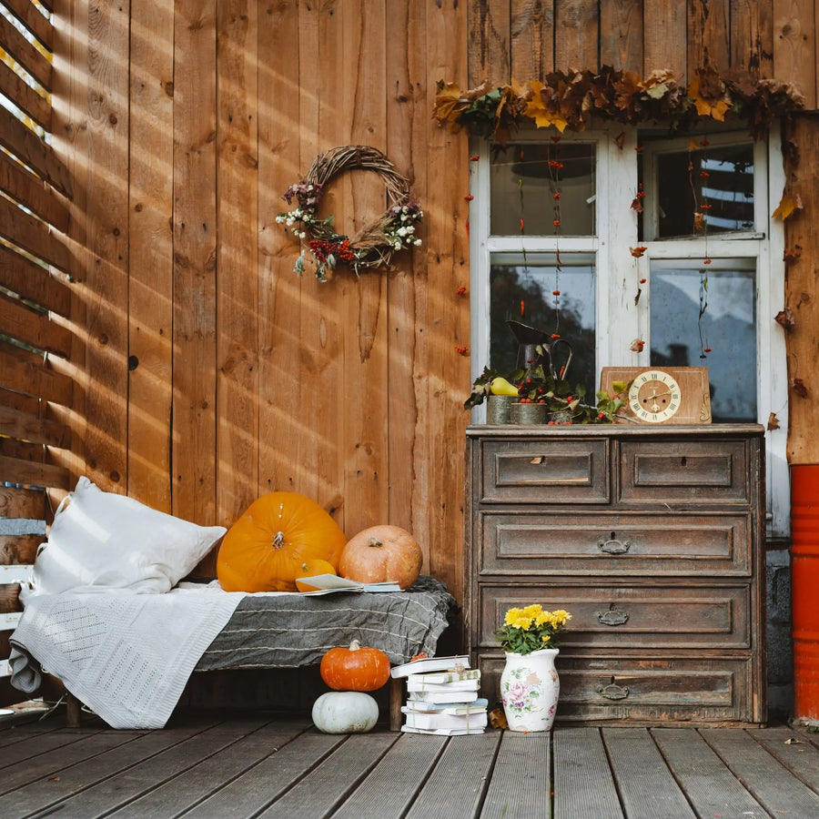 A rustic wooden porch decorated for fall with pumpkins, a wreath, a dresser, and a vase of yellow flowers.