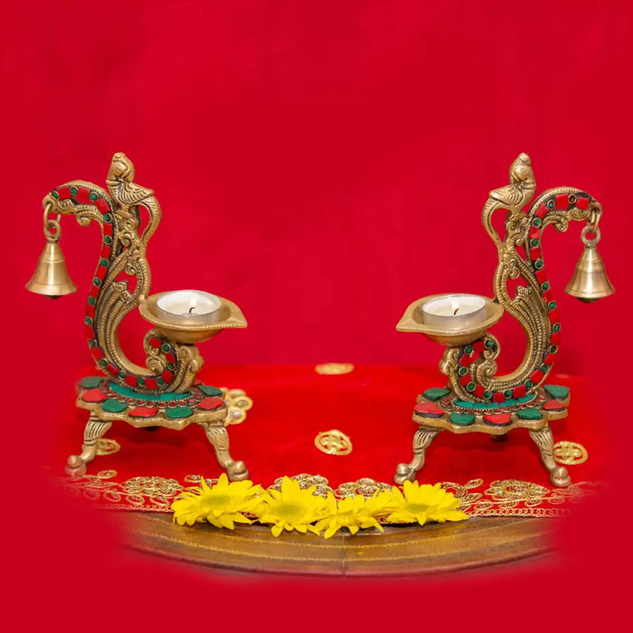 Two ornate brass diya holders with bells and lit tea lights sit on a red cloth with yellow flowers.