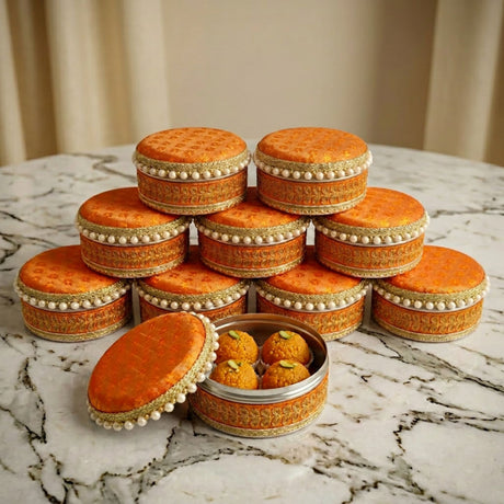 Ornate orange fabric-covered 4 ct steel ladoo boxes with gold embroidery and pearl trim, one open with golden sweets