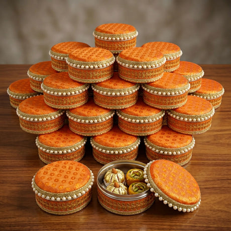 Ornate orange fabric covered round steel ladoo boxes with gold embroidery and pearl trim filled with golden sweets for festivals