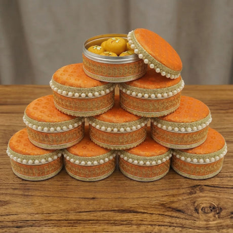 Orange fabric covered round steel ladoo boxes with gold embroidery and white bead trim stacked for festive gifting