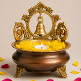 Golden ornate brass urli bowl with hanging bell and decorative handles featuring yellow marigold petals around lit candle