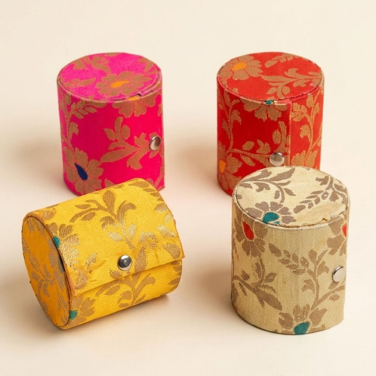 Four cylindrical fabric bangle box organizers in pink red yellow and beige with metallic floral patterns and snap closures