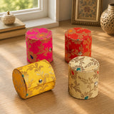 Four cylindrical fabric bangle boxes in pink red yellow and beige with gold floral patterns and snap closures for jewelry storage