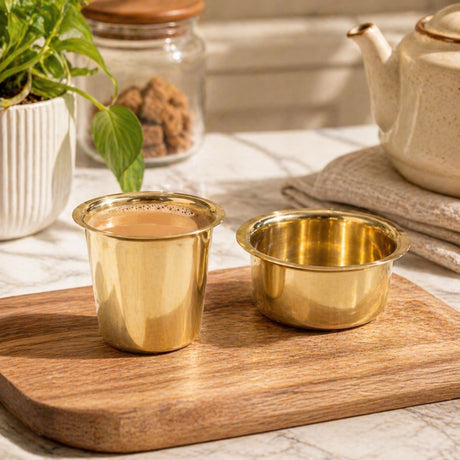 Two gleaming brass south indian coffee cups one frothy with tea the other empty on a rustic wooden board