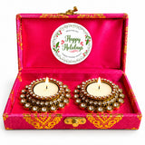 Two gold-plated diya lamps with clear crystals and lit candles in a pink satin box from happy holidays gift hamper