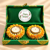 Happy holidays gift hamper with two ornate golden tealight candle holders adorned with yellow paper roses and white pearls inside a green satin-lined premium brocade box