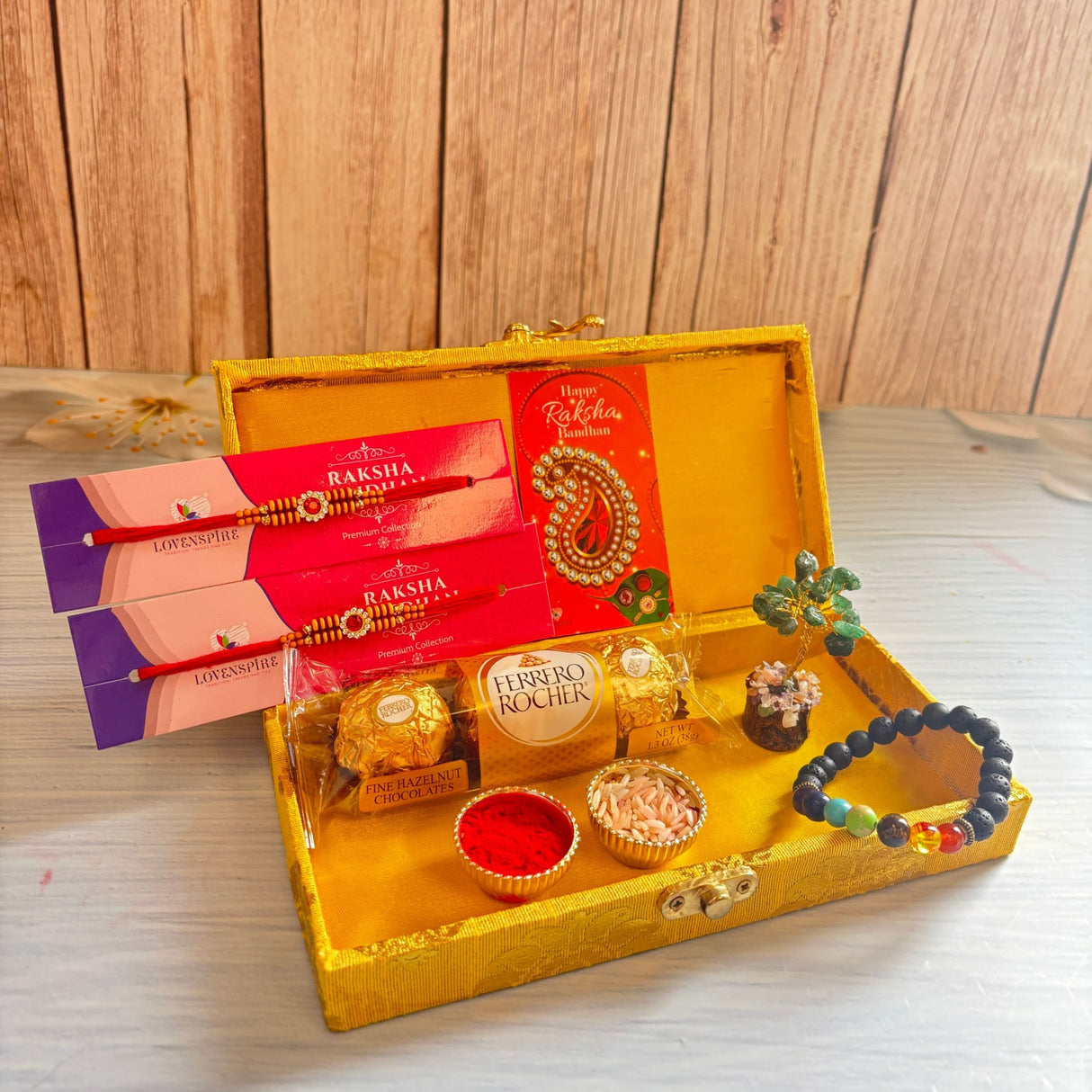 Golden ornate designer rakhi gift hamper with red and gold rakhis, chakra bracelet, chocolates, roli, and rice grains for brother and bhabhi