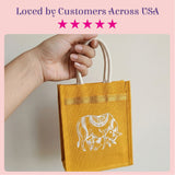 A mustard-yellow eco-friendly jute tote bag with white embroidered cow design and cream handles for Indian wedding favors