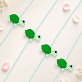 Green turtle-shaped charm with black and white eyes on light blue braided cord cartoon animal rakhi for kids
