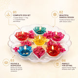A clear flower-shaped tray with seven colorful glass floating rangoli diyas with lit wicks and pink floral accents