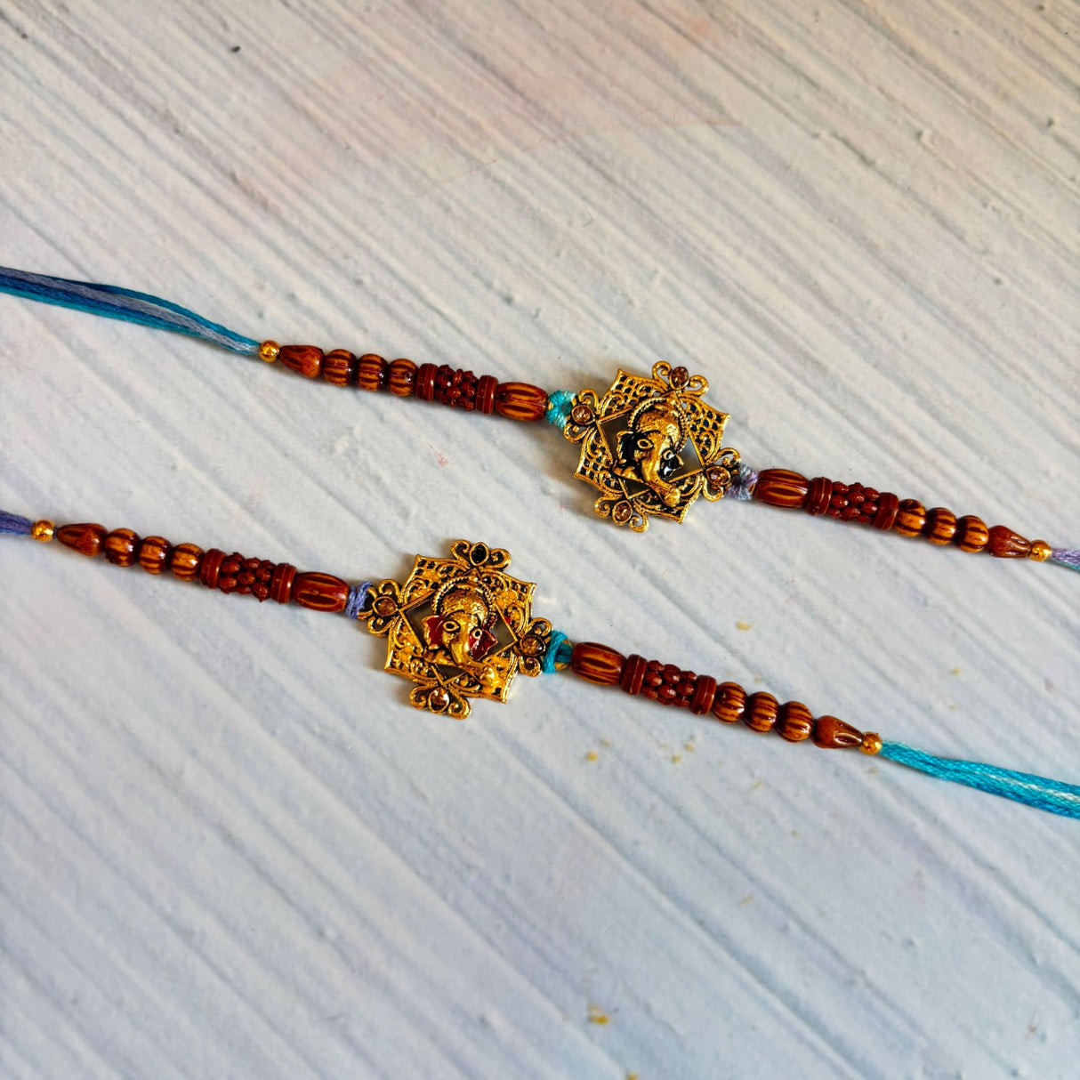 Two designer ganesha rakhi bracelets with golden pendants and reddish-brown wooden beads on turquoise cords for raksha bandhan