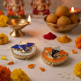 Two ornate gold-toned ganesha-themed diya holders with blue and orange enamel accents for diwali pooja rituals