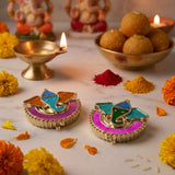 Two colorful glittery ganesha-themed diya holders with gold-toned metal bases and vibrant enamel accents for diwali pooja
