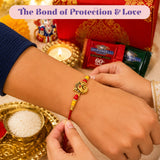 A golden ganesha-themed rakhi with red and yellow beads on a red thread from Raksha Bandhan gift hamper set