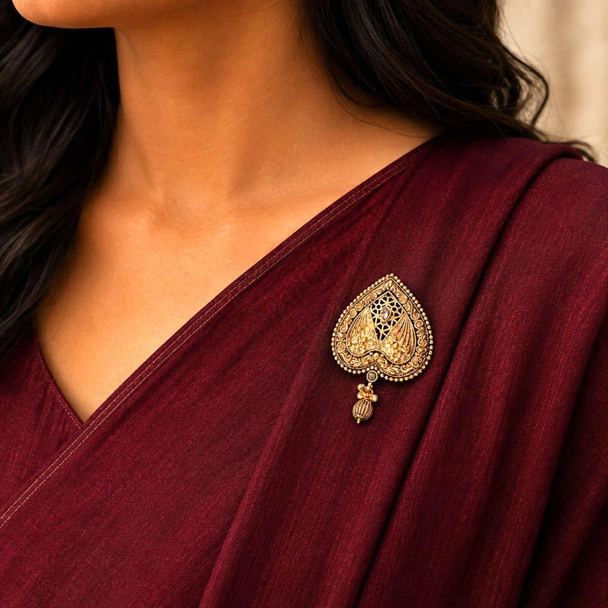 A gold-colored intricately filigreed heart-shaped sari brooch with a dangling bead accent for traditional Indian wear
