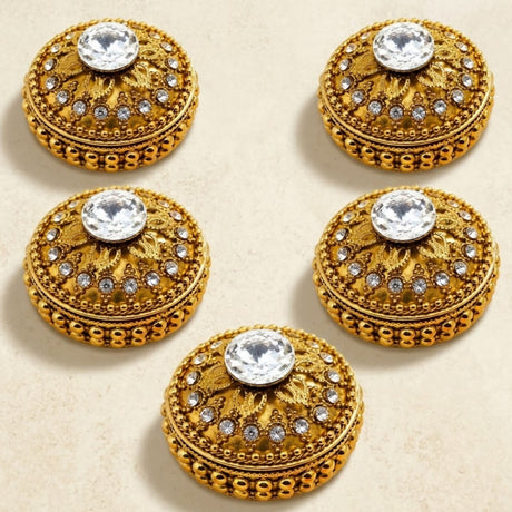 Gold-colored ornate button-like embellishments with intricate filigree and central clear crystal on golden kumkum holder