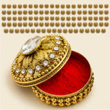 Golden metallic kumkum holder jewelry box with ornate design and clear gemstone lid featuring red velvet interior