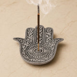 Silver german silver hamsa hand incense holder with intricate eye floral patterns holding a smoking stick for meditation