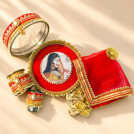 Red and gold embellished karwa chauth pooja thali set with circular mirror, pearl-trimmed containers, and decorative fabric