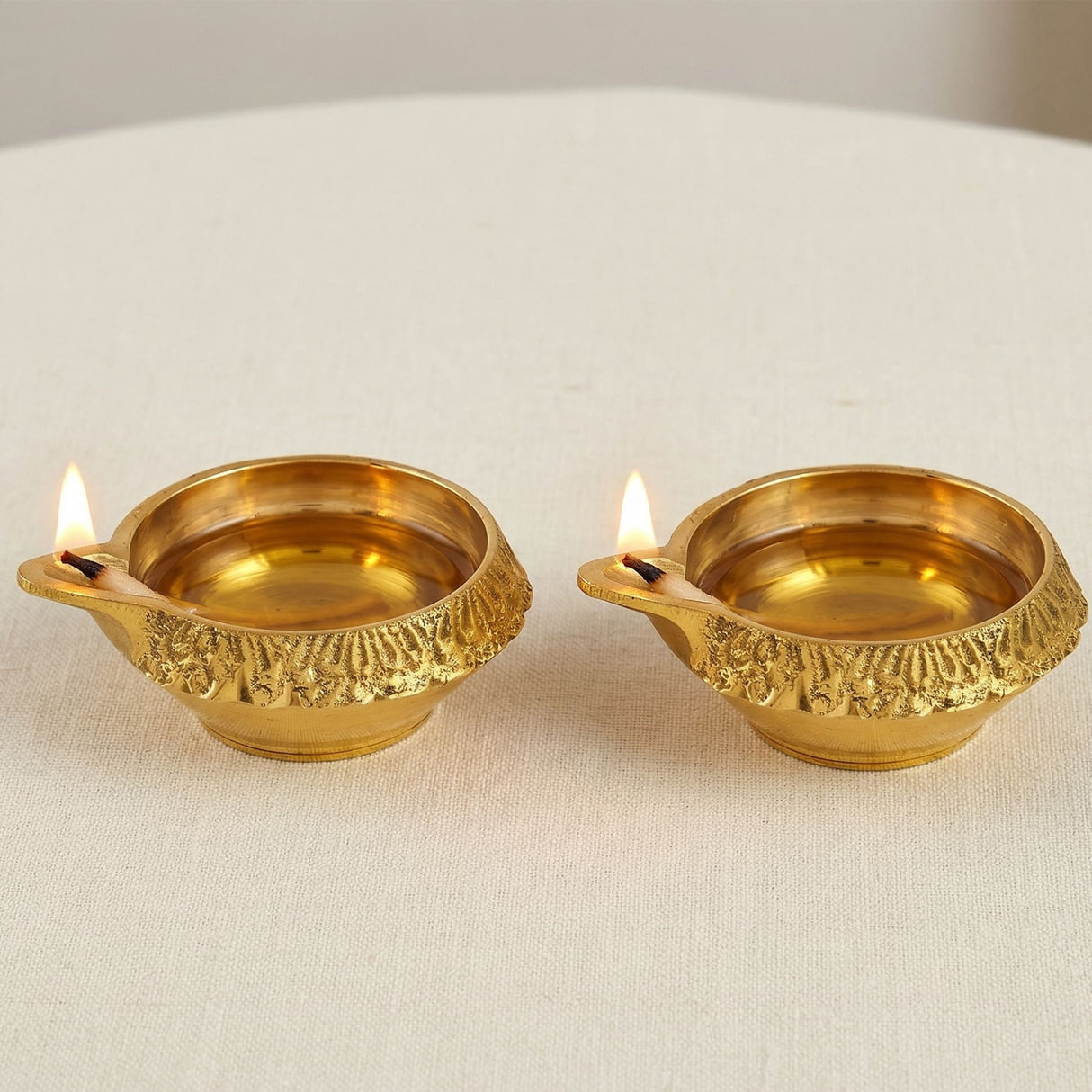 Two golden brass kuber diyas with intricate embossed patterns holding lit wicks for diwali and puja decor