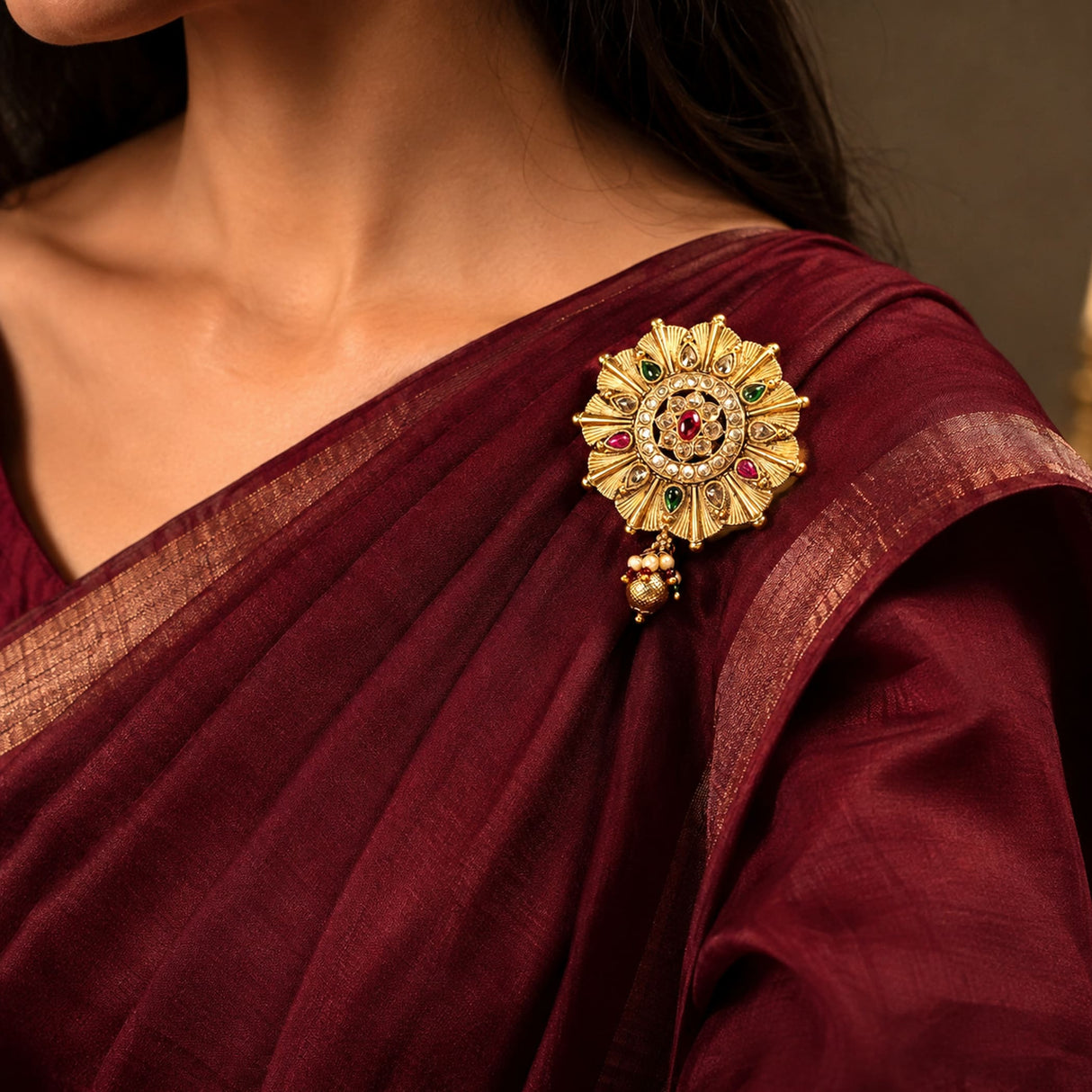 Gold-colored intricate kundan sari brooch with red and green gemstones and a dangling pearl charm for bridal wear