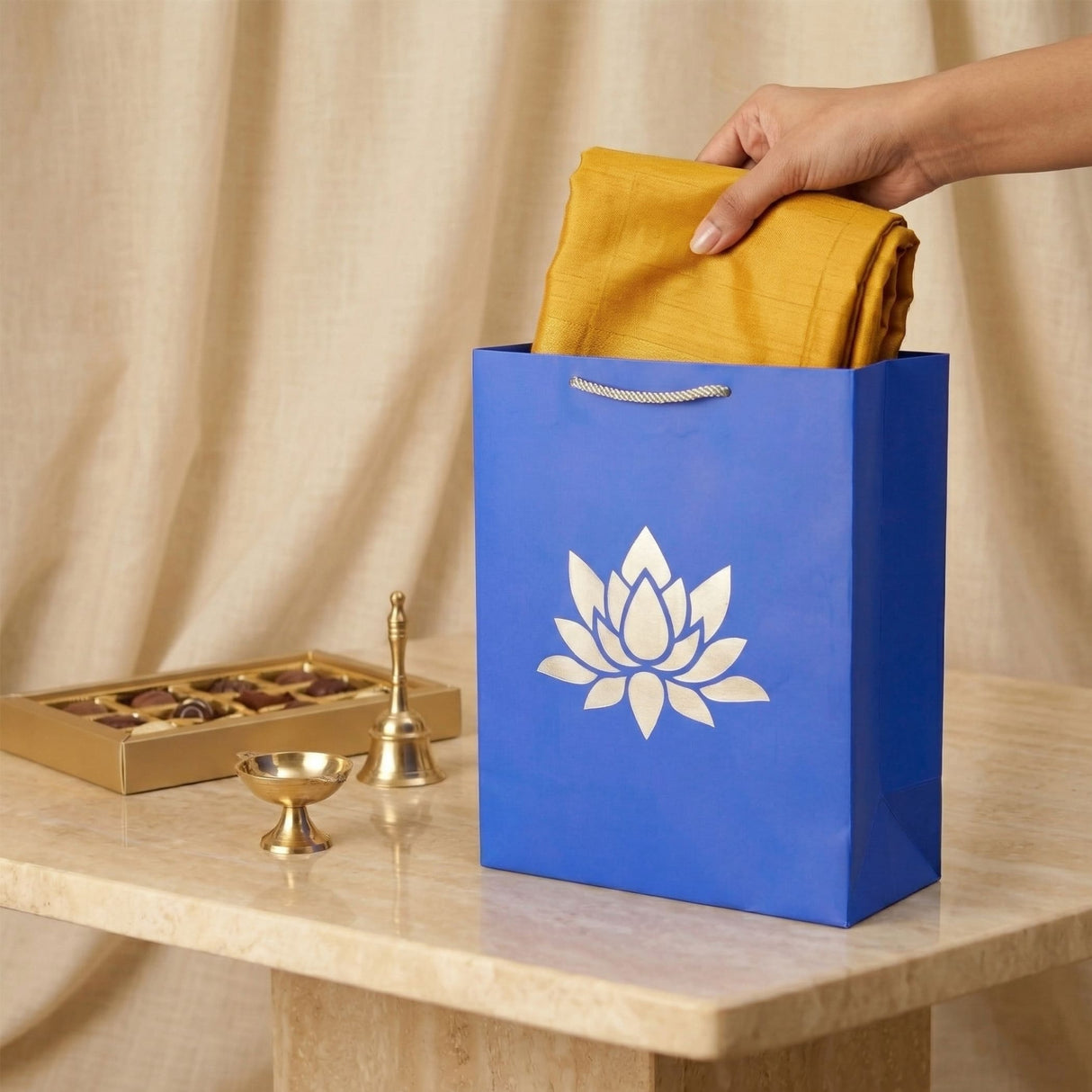 Blue paper gift bag with white lotus logo and silver handle holding mustard-yellow fabric for Indian gift occasions