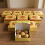 Yellow cardboard sweet box with ornate red and green mandala design containing golden brown round sweets for gifting