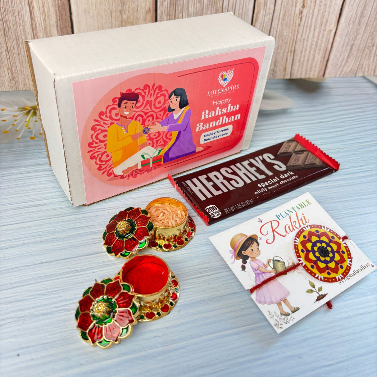 A vibrant red and gold floral enamel diya with matching saucer in designer mandala seed rakhi gift hamper for brother