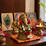 Golden ganesha statue adorned with red and white artificial flower garlands seated on red and gold cushion for diwali puja