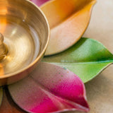 A golden brass oil lamp with a smooth surface sits on colorful ceramic leaf holders for traditional Diwali puja