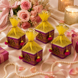 Purple and gold ornate small potli gift boxes with yellow organza drawstrings and pink pom-pom tassels for festive use