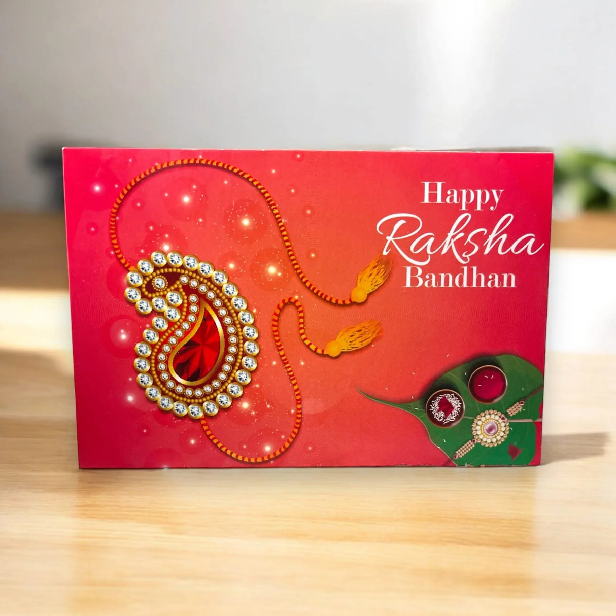 Red greeting card with gold silver paisley design and orange tassels alongside green rakhi with red gemstones for raksha bandhan