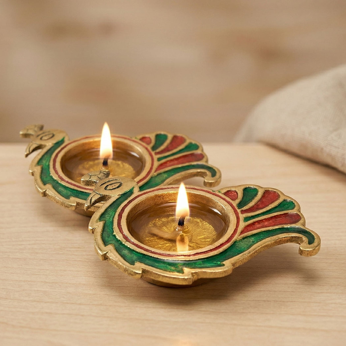 Two ornate gold-toned brass peacock-shaped diya lamps with green and red enamel holding lit candles for diwali puja