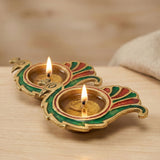 Two ornate gold-toned brass peacock-shaped diya lamps with green and red enamel holding lit candles for diwali puja