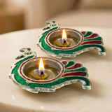 Two ornate peacock-shaped brass finish diya lamps with silver base and green red enamel for Diwali Puja decor